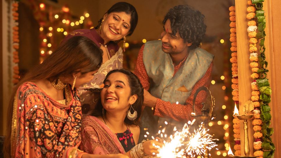 A family celebrating Diwali at home, the house is decorated with garland and two young women are using sparklers as a young man and a mother watch.