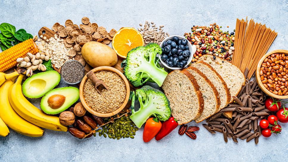 An assortment of foods providing fiber, including bananas, broccoli, avocado, corn, beans, whole grain bread, whole grain pasta, tomatoes, peppers, nuts and blueberries.