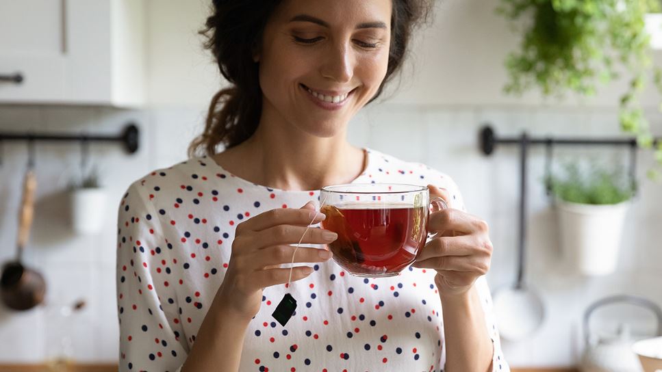Woman holding a glass mug, standing in kitchen, smelling and drinking hot tea beverage