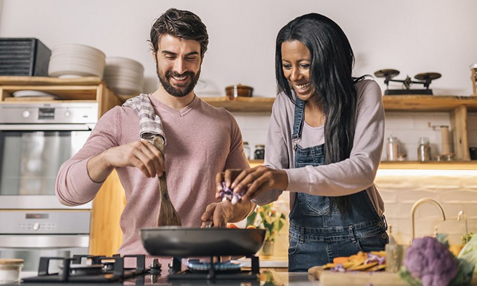 A couple smiling and cooking food together in the kitchen at home.