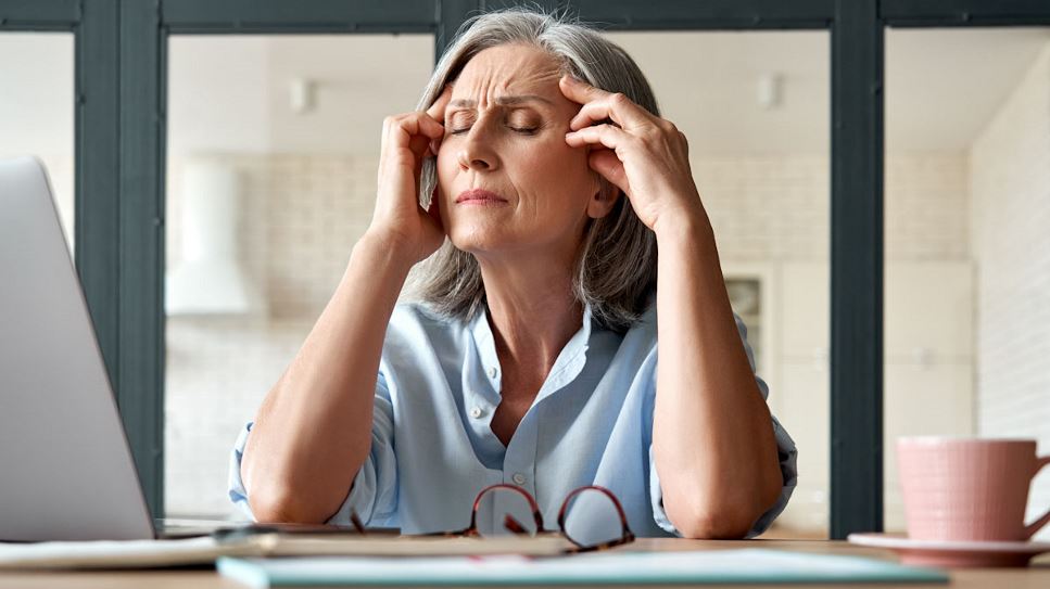 Woman with grey hair sits at computer desk with her eyes closed and hands on her forehead.