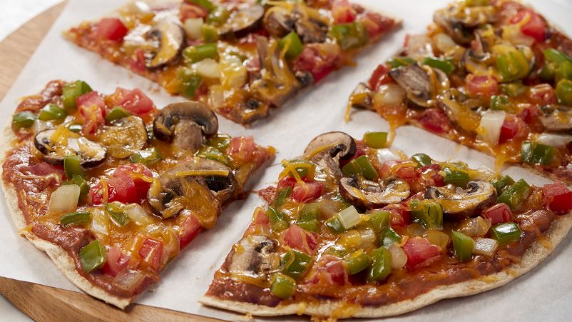 Four pieces of sliced pieces with mushrooms, peppers and tomatoes on a piece of paper that's on a round wooden slab.