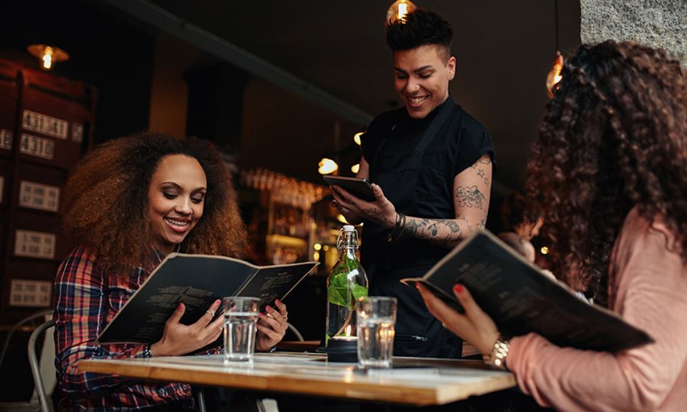 Two young women are at a trendy cafe ordering from restaurant menus that feature sustainability terms and phrases.